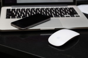 An iPhone, laptop and mouse sitting on a black desk.